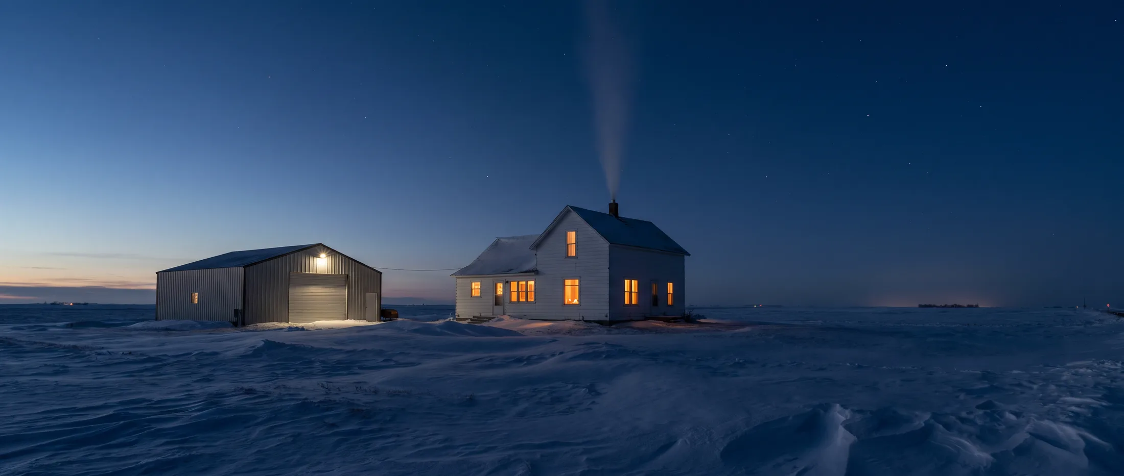 A farmhouse and shop glowing warm at dusk on the snowy North Dakota prairie